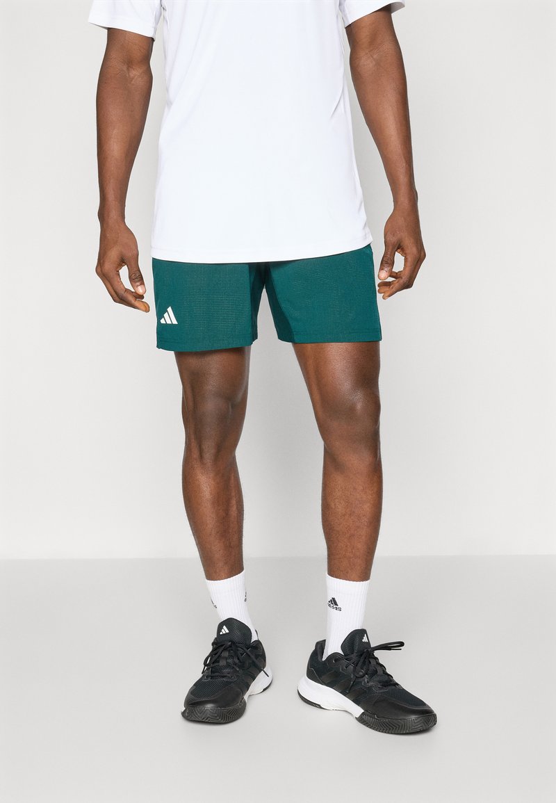 Shorts de sport vert foncé avec un motif texturé, arborant un logo blanc sur la jambe gauche. Associés à un t-shirt blanc et des baskets noires.