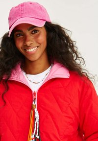 Červená prešívaná bunda s ružovým golierom, bielou košeľou pod ňou a ružovou čiapkou. Obsahuje zips s farebnými šnúrkami a náhrdelník z korálikov.