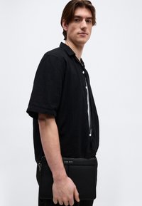 Νέος άνδρας με μαύρο πουκάμισο με κοντά μανίκια κρατώντας μια μαύρη τσάντα με φερμουάρ μπροστά σε απλό ανοιχτόχρωμο φόντο.