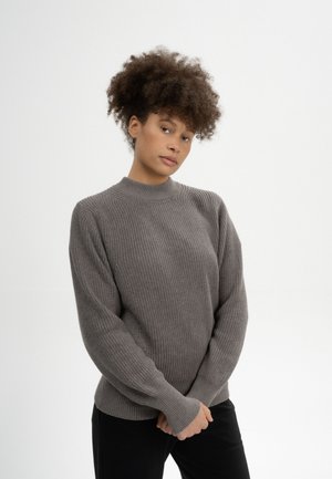 Vrouw met krullend haar die een grijze gebreide trui en zwarte broek draagt, staand tegen een witte achtergrond met gevouwen handen.