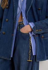 Χέρι στην τσέπη με σκουρόχρωμο τζιν μπουφάν και τζιν παντελόνι με χρυσή αλυσίδα, συνδυασμένα με μπλε και λευκό ριγέ πουκάμισο.