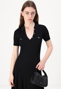 Black ribbed dress with a V-neck, short sleeves, and silver-button accents. Model holds a small black handbag. Plain white background.