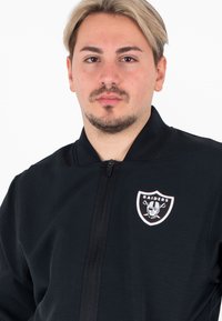 Giacca nera con chiusura zip frontale, colletto a coste e logo dei Raiders sul lato sinistro del petto. Tessuto morbido con una consistenza liscia e un design minimale.