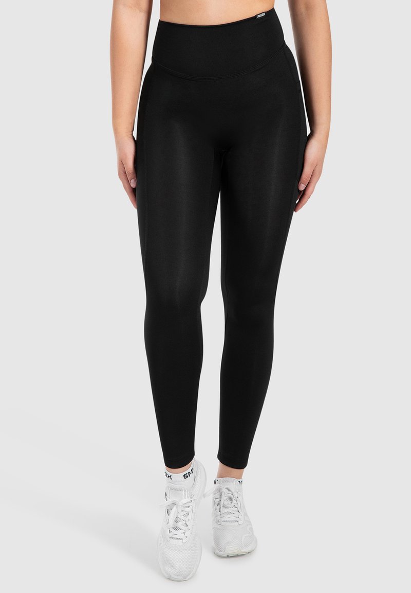 Leggings noirs taille haute en tissu extensible et lisse. Présente un design épuré et épouse les jambes de près. Portés avec des baskets blanches.