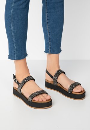 Fødder iført sorte platformsandaler med glitrende remme parret med cropped blå denim jeans på hvid baggrund.