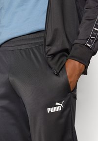 Črne trenirke z gladko teksturo, z belim logotipom Puma in stranskim žepom. V kombinaciji z svetlo modrim topom pod njimi.