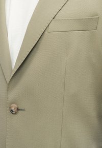 Σακάκι ταμπά με πέτο notch, ένα καφέ κουμπί και θήκη στήθους, φορεμένο πάνω από λευκό πουκάμισο.