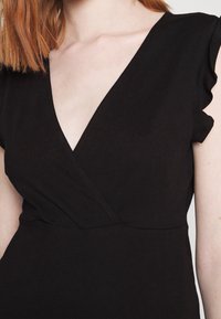 Robe noire avec un col en V et des manches volantées, confectionnée dans un tissu doux à la texture lisse avec des détails de coutures visibles.