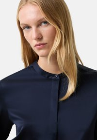 Marineblauwe blouse met knopen gemaakt van gladde stof met een mandarijnkraag en subtiele glans. Heeft een enkele knoopsluiting bij de hals.