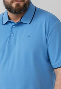 Lichtblauwe polo van gestructureerde stof, met een donkerblauwe kraag en een klein logo op de borst, voorzien van een tweeknops knoopsluiting.