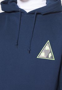Sudadera con capucha azul marino con cordones, que presenta un gráfico triangular en verde claro y rayas estampadas en crema con las letras "huf."