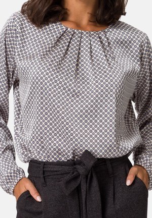 Femme portant un chemisier blanc avec de petits motifs circulaires noirs et un pantalon gris foncé taille haute avec une ceinture en tissu nouée.
