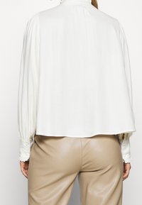 Weiße, locker sitzende Bluse mit plissierten Ärmeln und hohem Kragen; kombiniert mit enganliegenden beigen Hosen. Glattes Material mit dezentem Glanz.