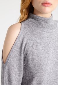 Pull gris tricoté avec un col roulé et des découpes ouvertes aux épaules. Le tissu a une texture douce et une épaisseur moyenne.