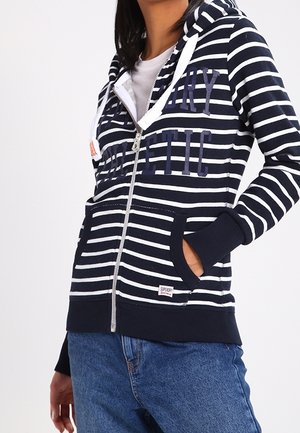 Kvinde iført marineblå og hvide stribede lynlåshættetrøje med forlommer og broderet tekst, kombineret med blå jeans, stående mod en hvid baggrund.