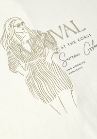 Chemise en tissu blanc avec un dessin doré représentant une silhouette en tenue rayée. Le texte inclut "ARRIVÉE À LA CÔTE" dans une police élégante.