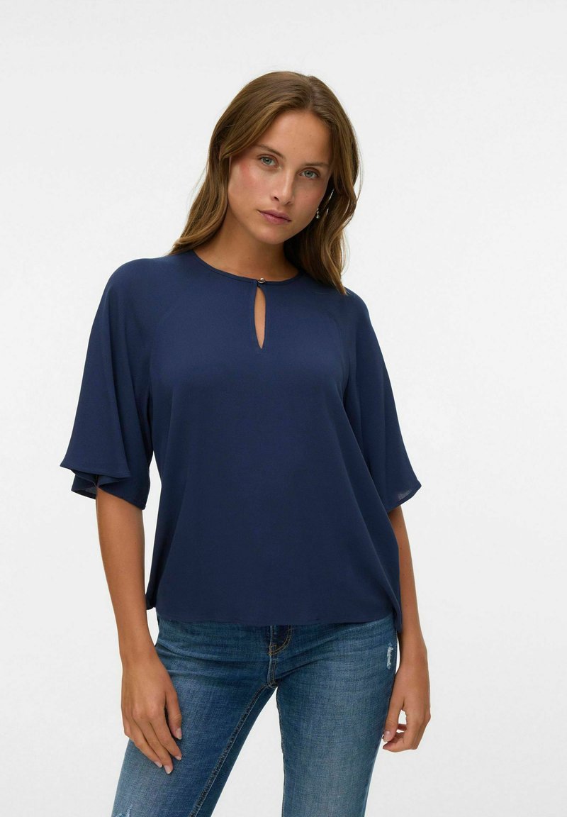 Donna con capelli castano chiaro che indossa una blusa blu navy con scollatura a chiave e maniche a campana, abbinata a jeans blu su uno sfondo bianco.