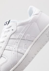 Bílá kožená teniska s texturovaným svrškem, kulatou špičkou a perforovanými detaily. Měkké vnitřní podšívání a plochá gumová podrážka. Logo značky na patě.