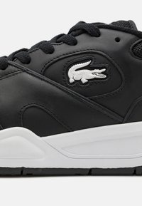 Μαύρο δερμάτινο αθλητικό παπούτσι με λευκή καουτσούκ σόλα, υφή στις λεπτομέρειες, μαύρες κορδόνες και λευκό λογότυπο κροκοδείλου στο πλάι.