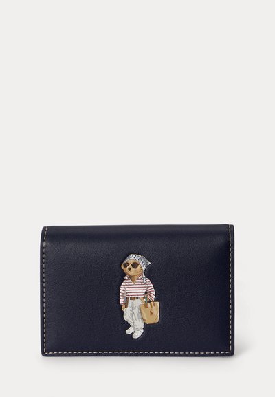 Πορτοφόλι από ναυτικό δέρμα με ραμμένες άκρες, που απεικονίζει μια αρκούδα με γυαλιά ηλίου, ριγέ πουκάμισο, λευκό παντελόνι, μαντήλι στο κεφάλι και που κρατά τσάντα tote.