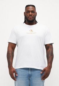 Baltas medvilnės marškin�ėliai su auksiniu "CK" logotipu ir "Calvin Klein Jeans" tekstu ant krūtinės. Trumpomis rankovėmis ir paslėgto silueto.