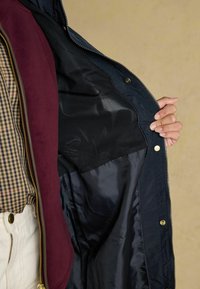 Jachetă marină deschisă pentru a dezvălui o căptușeală din plasă neagră. Exteriorul are un fermoar auriu și nasturi, asociate cu un accent de guler în culoare vișinie.