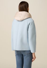 Žena viděná zezadu, která má na sobě světle modrou bundu s béžovou prošívanou kapucí a modré džíny na jednobarevném béžovém pozadí.