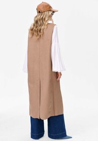 Beżowa długa kamizelka o prostym kroju, z bocznymi rozcięciami i pikowanym wykończeniem, noszona na białej bluzce i zestawiona z dżinsowymi spodniami o szerokich nogawkach.