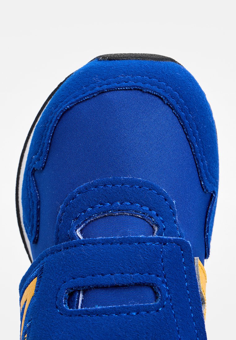 Chaussure de sport bleue avec une tige en daim et nylon, bande Velcro, bout arrondi et semelle en caoutchouc noir. Accentuation jaune notable sur le côté.