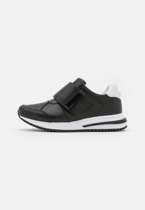 Baskets noir et blanc avec semelle blanche épaisse et large fermeture Velcro, vue de côté sur fond clair uni.