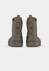 Et par høye, brune vanntette støvler med trekkremmer og robuste Vibram-såler, vist fra baksiden på en hvit bakgrunn.