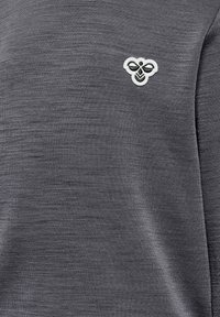 Grå stickad topp med en liten broderad bi-logotyp i vitt och grönt på vänster bröst. Texturerad tyg med en slät finish.