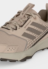 Μπεζ παπούτσι για τρέξιμο σε μονοπάτια Adidas Terrex με υφή υφάσματος, ελαστική σόλα και τρεις γκρι ρίγες στη πλευρά.