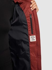 Bordeauxrote Jacke mit glänzendem marineblauem Innenfutter, ausgestattet mit einer Reißverschlusstasche und einem Etikett mit der Aufschrift "Eine warme Umarmung aus Stoff."