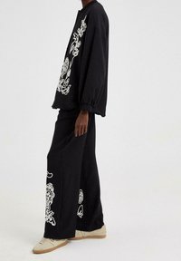 Schwarzes Baumwoll-Outfit mit einem lockeren Sweatshirt, das mit weißen Blumenstickereien verziert ist, und passenden weit geschnittenen Hosen, ergänzt durch beigefarbene Sneakers.