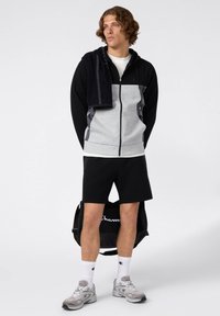 Graue und schwarze Reißverschluss-Hoodie, schwarze Shorts, graue Turnschuhe, weiße Socken mit Logo. Handtuch über der Schulter, trägt eine schwarze Tasche.