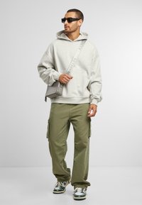 Lichtgrijze hoodie met kangoeroezak, olijfgroene cargobroek met zijzakken en zwarte zonnebril. Grijze schoudertas met strapdetails.