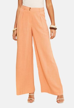 Vrouw die perzikkleurige hoge taille broek met wijde pijpen en dubbele knoopsluiting draagt, witte top, sandalen met koordriempjes en opvallende armbanden aan beide polsen.