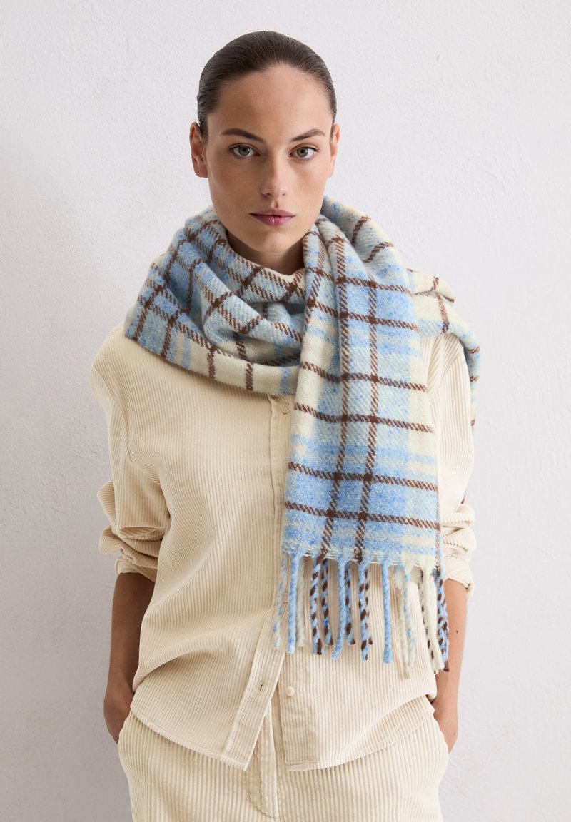 Wollschal mit einem blau-cremefarbenen Karomuster und braunen Linien, Fransenenden, getragen über einem hellfarbenen, gerippten Hemd.