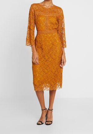 Koronowa sukienka w kolorze rdzawego pomarańczu sięgająca do kolan, z rękawami do łokcia, dopasowaną talią i kwiatowymi wzorami, zestawiona z czarnymi szpilkami na kostkę z paskiem.