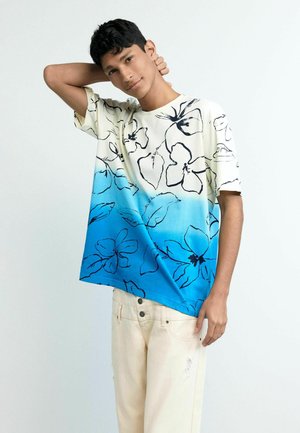 Młody mężczyzna pozuje z ręką za szyją, ubrany w białą i niebieską koszulkę z gradientem oraz czarnymi kwiatowymi konturami i jasnobeżowe spodnie.