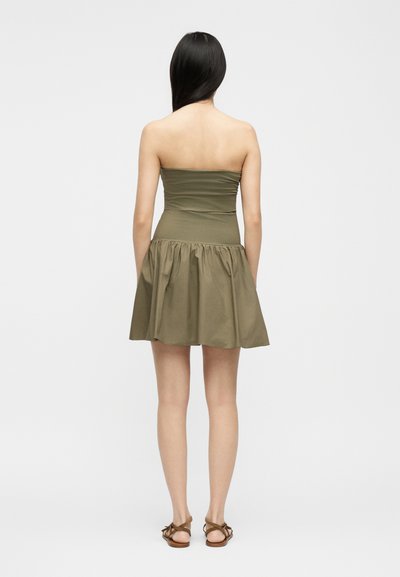 Γυναίκα με μακριά σκουρόχρωμα μαλλιά, φορόντας ένα χακί φόρεμα χωρίς τιράντες και καφέ σανδάλια, στέκεται γυρισμένη πλάτη σε λευκό φόντο.