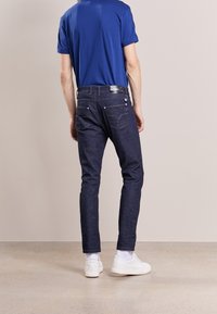 Modrá polokošile s krátkým rukávem, tmavě modré slim-fit džíny se zadními kapsami a bílé tenisky, zobrazené ze zadní strany na neutrálním pozadí.