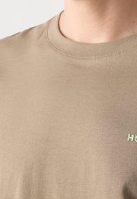 Nærbillede af en person iført en beige T-shirt med rund hals, hvor delvis grøn tekst "HU" er synlig på højre side.