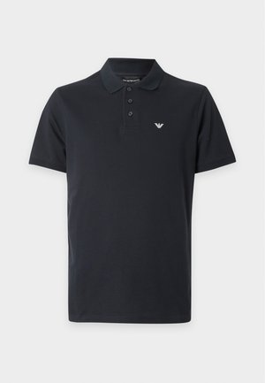 Uomo con polo nera, pantaloncini beige, calzini neri e scarpe, che porta una borsa a tracolla con motivo e cinghia scura sulla spalla.