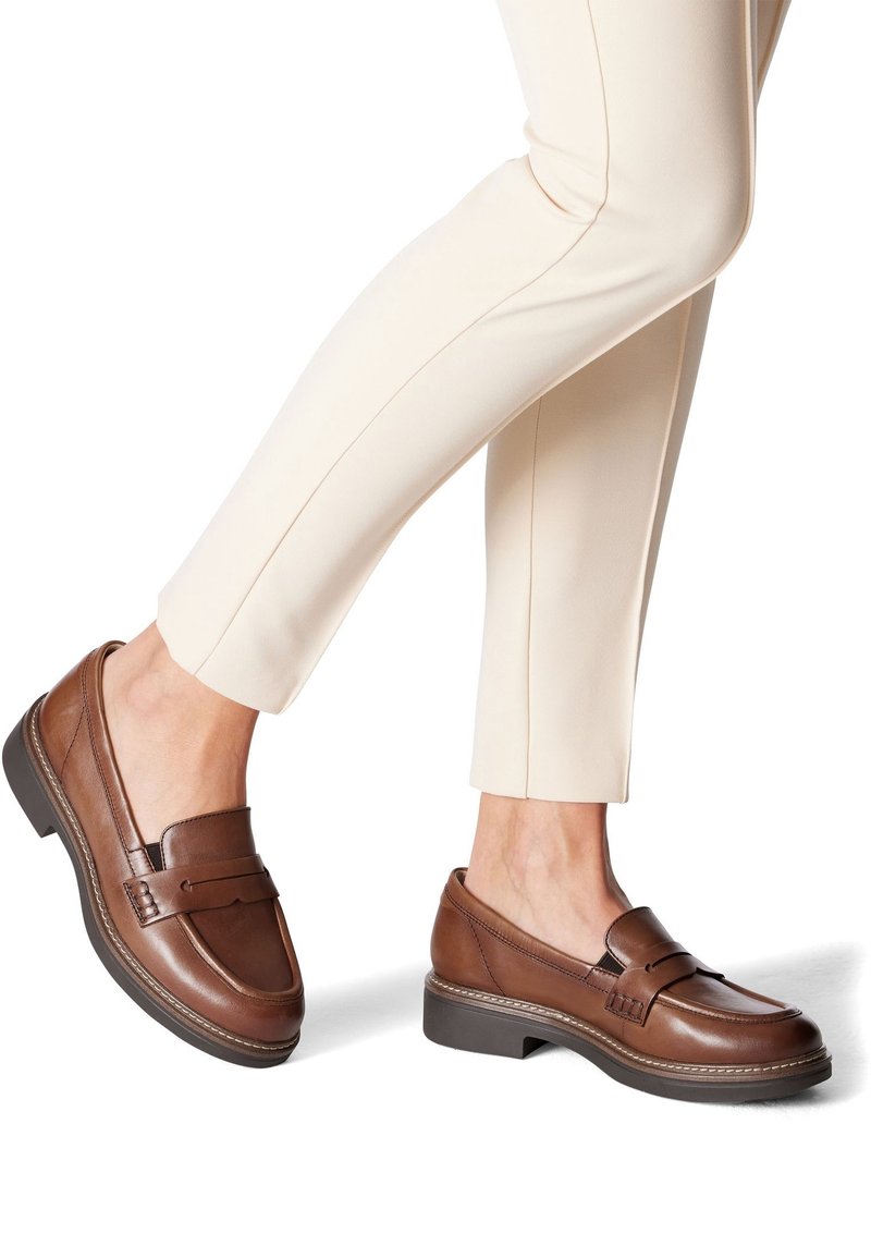 Personne portant un pantalon beige et des mocassins en cuir marron avec des semelles noires, debout avec un pied légèrement levé sur un fond blanc.