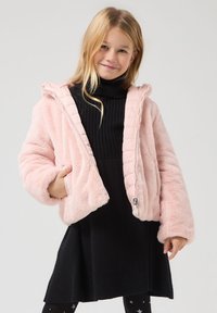 Giacca in pelliccia sintetica rosa con cappuccio, zip frontale e maniche testurizzate, abbinata a un vestito nero a girocollo a coste e collant fantasia.