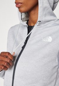 Felpa grigia con zip, dotata di zip nera e coulisse, con il logo The North Face. Texture liscia e design aderente.