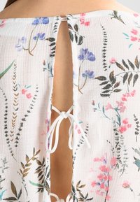 Tessuto bianco con motivo floreale caratterizzato da fiori viola e rosa. Design con schiena aperta e chiusura a cordoncino bianco annodato.