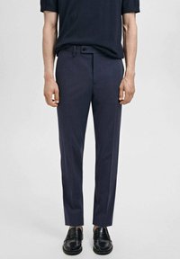 Pantalons taillés en marine en tissu lisse, présentant une coupe ajustée, des passants de ceinture et une fermeture à un bouton. Associés à des chaussures noires.